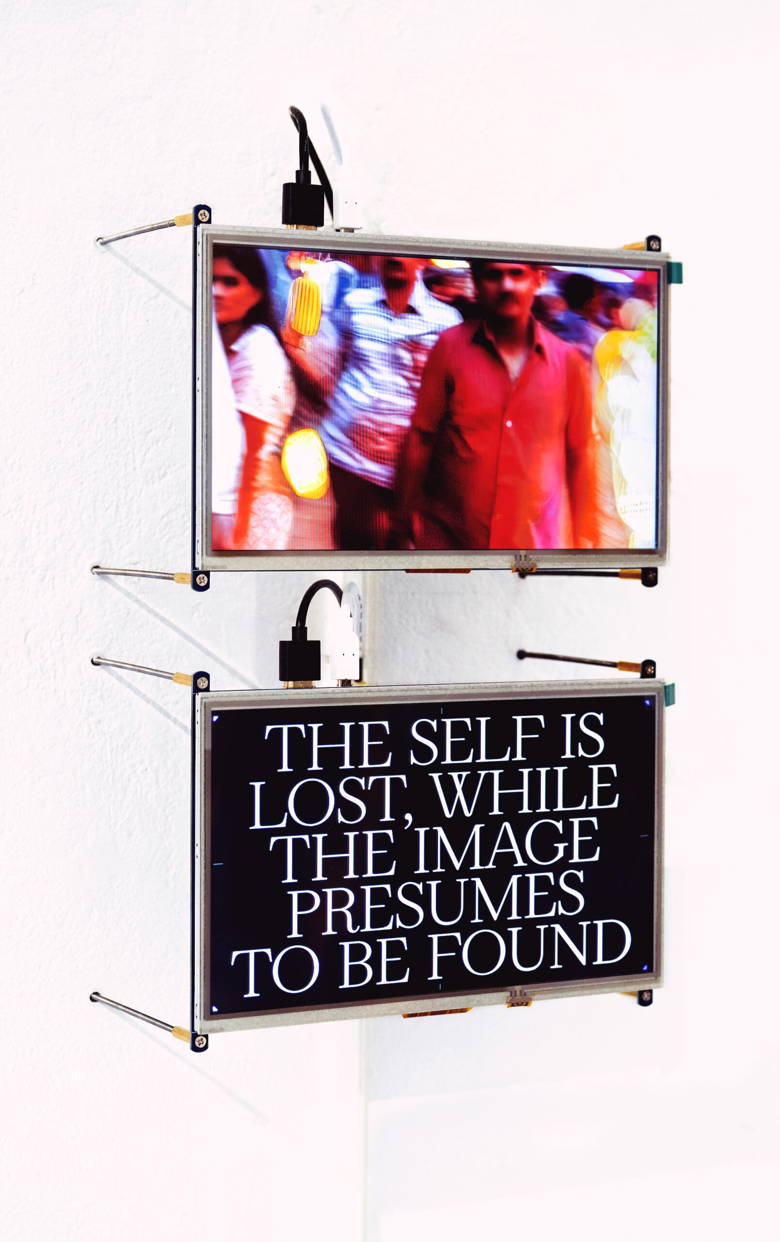 A photograph of a two-channel video installation, with two thin small-sized screens placed vertically on a wall. The screen above shows an video of crowds of people, while the screen below depicts a text that reads "The self is lost, while the image presumes to be found". This text is in styled in all capitals, white serif font on a black background.